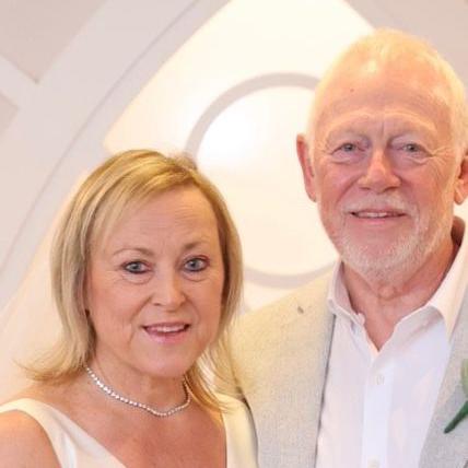 Anita Fox is pictured smiling into the camera on her wedding day, alongside her husband, Paul. She is wearing a cream satin V neck dress and a pearl necklace. Her hair is blonde and shoulder length. Paul stands next to her, wearing a white shirt and grey suit with a buttonhole. He has short, grey hair.