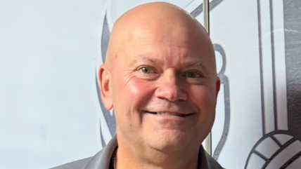 A bald man with blue eyes, wearing a grey top, smiles at the camera.