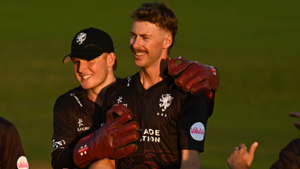 Thomas Rew celebrates with Riley Meredith, with both players wearing Somerset's black shirt. Rew has his arms around Meredith and both are smiling.