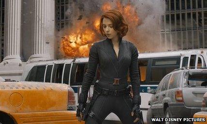 Scarlett Johansson as Black Widow stands in front of an exploding vehicle.