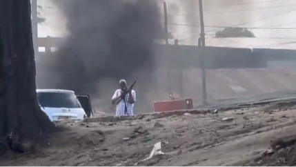A man in civilian clothes holds a rifle, standing beside a saloon car. Black smoke rises behind him.