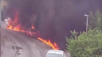 A strip of flames runs down a road. In the centre, you can se a small red car with a black cross on its front. Lots of black smoke blocks the view behind the road.