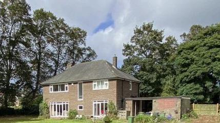 A two‑storey brick house situated in a large, open garden area bordered by tall, mature trees. The house has a dark, hipped roof and several white-framed windows on both the ground and upper floors. A small extension or outbuilding is attached to the right-hand side of the property, constructed from similar brickwork but with a flatter roof. In front of this structure, there are a few items placed on the ground, including garden equipment and containers.