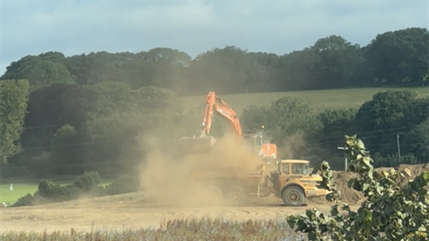 A construction site in a rural setting where machinery with a bright orange arm is dumping dirt into a large yellow dump truck. There is clouds of dust in the air. There is a green fields and trees in the background.