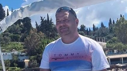 A picture of Tomasz Kopec, who is wearing a white, red and blue Tommy Hilfiger T-shirt, with trees and buildings in the background. He has sunglasses resting on the top of his head.