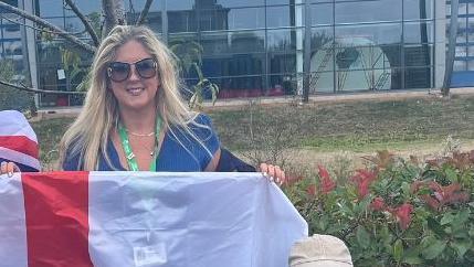 Alison Sheridan wearing sunglasses and a green Exeter City Council lanyard and a blue dress and holding a flag of St George's cross