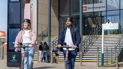 A woman with blond hair, wearing a pink coat and blue jeans, and a man with a dark beard wearing a blue jacket and a white shirt over blue trousers, ride pink e-scooters.  Both are wearing helmets.  They are in front of the steps which lead to the entrance of the modern glass-fronted Northampton station.