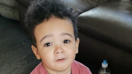A photo of Daniel Aruebose. He is a young child, around two and a half years old, who is standing near a brown leather sofa. He is wearing a long-sleeved red top and is looking at the camera.