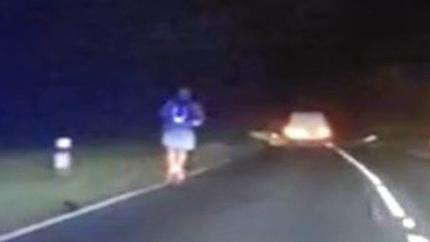 The e-scooter rider travelling on the A-road wearing a purple T-shirt and grey shorts. He is on the left of the road at night time and it is dark. A car is being driven in front of him. The picture appears to be a still taken from dashcam footage.