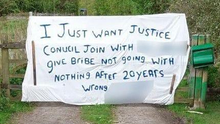 A white sheet is attached to a gate with a green letter box attached to one post. It has writing on it which starts with "I just want justice" at the top, followed by 'Council join with give bribe not going with nothing after 20 years wrong'. There is a track leading up to the gate with grass growing in the middle.