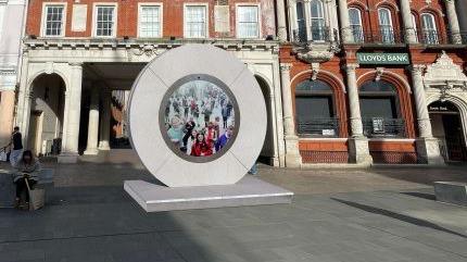 A computer generated image of what the portal could look like. It is a grey circle with a screen in the middle, it is on the Cornhill in Ipswich with buildings in the background. 