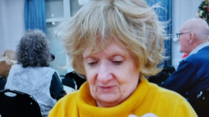 Woman with blonde hair looking down. She wears a yellow roll-neck jumper. There are people seated behind her at tables.