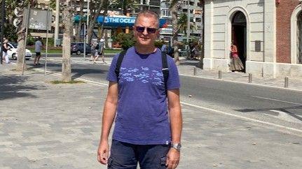 Chris Howard has short hair and is wearing a purple t-shirt and sunglasses. The sun is shining and he appears to be abroad.