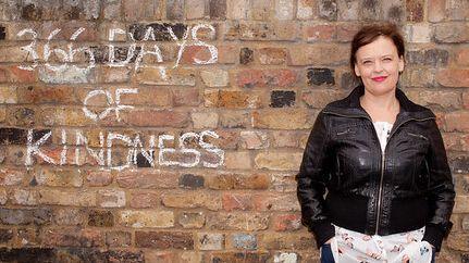 A woman stands in front of brick wall with '366 DAYS OF KINDNESS' written on it in white 