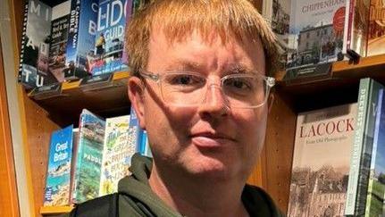 Chris stands smiling at the camera in front of a bookcase. He has ginger hair and is wearing clear rim glasses and a green hoodie.