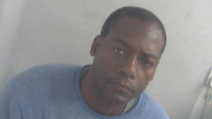 Errol McCardo Weir is in the middle of the image looking towards the camera. He has short black hair and is wearing a grey top with a blue jumper over the top. He is sat in front of a grey background.