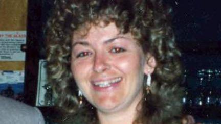 Deborah Steel is smiling at the camera. She has curly shoulder length hair and earrings. The camera flash has put a red shine in the pupils of her eyes.