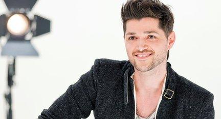 Danny O'Donoghue in a publicity photograph for BBC The Voice. He sits in a studio smiling, wearing a smart, trendy black jacket.