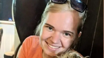 Beth Matthews, with blonde hair and wearing an orange t-shirt, smiles at the camera