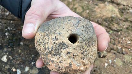 A hand holding what appears to be a Roman archaeological find. The artefact is round with a small hole going through the middle.