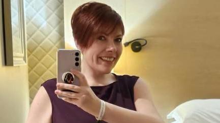 Kat Watson-Wood with short brown hair dressed in a burgundy evening dress in a hotel room holding her phone up to take a selfie. She is smiling.
