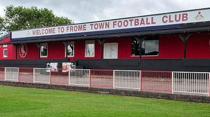 A photo of Badgers Hill stadium, the grass pitch and goal in front of a covered terrace with the words 'Welcome to Frome Town Football Club'