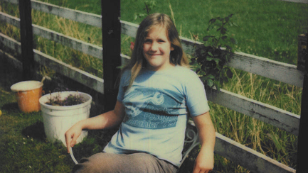 A photograph from the 1970s of a young girl wearing a blue T-shirt and trousers as she sits in the garden. There are plant pots and a wooden fence bordering a field behind her.