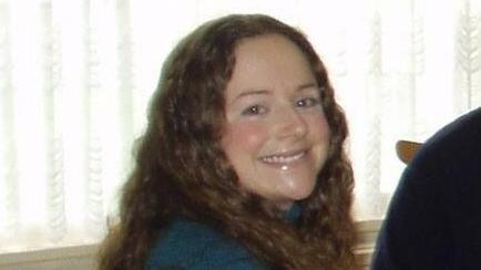 A young women with brown wavy hair is looking at the camera and smiling.  She is wearing a blue jumper