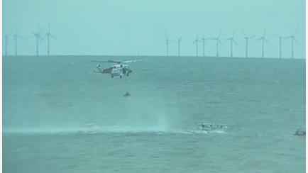 A helicopter hovering over the sea and winching someone up. There are wind turbines in the background.