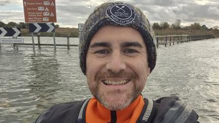 Oli Jordan has a beard, he is in a kayak and taking a selfie in front of road signs while smiling at the camera. He is wearing a beanie hat and a coat. 