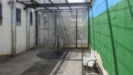 An outdoor recreation area of a prison enclosed by high walls and a ceiling, with a concrete floor and a piece of metal exercise equipment