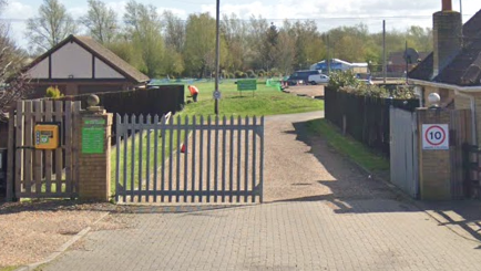 Two silver gates are in between two houses. The one on the right is open and a driveway leads to a building. There is a 10mph sign on a pillar on the right and a defibrillator is on the left gate.