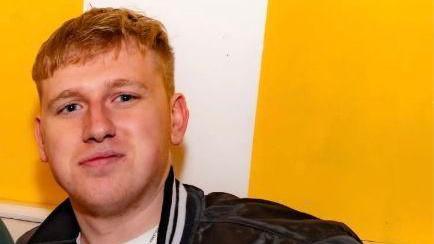 A man with short ginger hair smiling and looking at the camera. He is wearing a black jacket and white top. He is standing in front of a white and yellow wall.