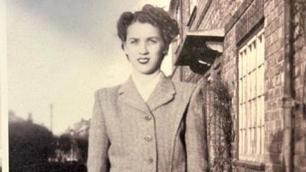 A black and white photo of a woman with short curly hair, wearing a suit jacket. She stands on a street with, to her left, a house with a latticed window. Behind her look to be further homes in the distance.