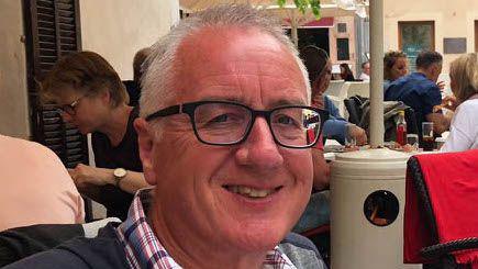 Gordon Robb has short, grey hair and black glasses and is wearing a checked shirt. He is smiling and sitting at an outdoor restaurant with people sitting at tables behind him.