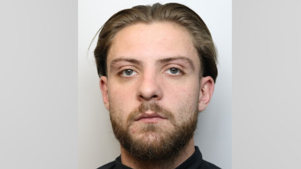 Mugshot of a man with brown slicked back hair and a beard. He looks into the camera with a serious expression.