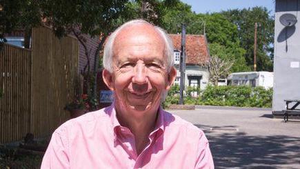 Neil Watts smiles at the camera while standing outside. He wears a pink shirt and has grey hair.