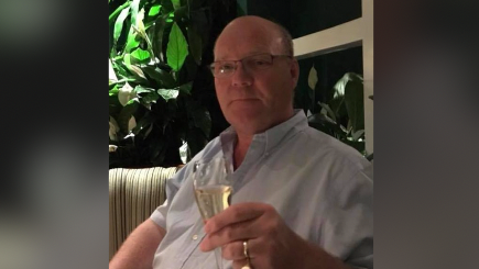 Bald haired man in a blue shirt. He's got his top button unbuttoned. He's wearing glasses and looking at the camera. He's holding a champagne glass in in his right hand.