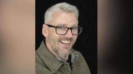 Craig Hurcombe smiles at the camera. He wears a green collared jacket and black rimmed glasses. He has short grey hair and a beard.