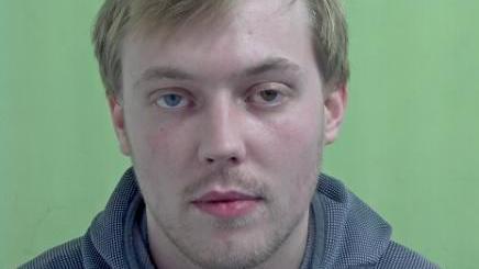 Police mugshot of Lewis Kelly, who has short blonde hair and is wearing a grey hooded top. The background behind him is green.