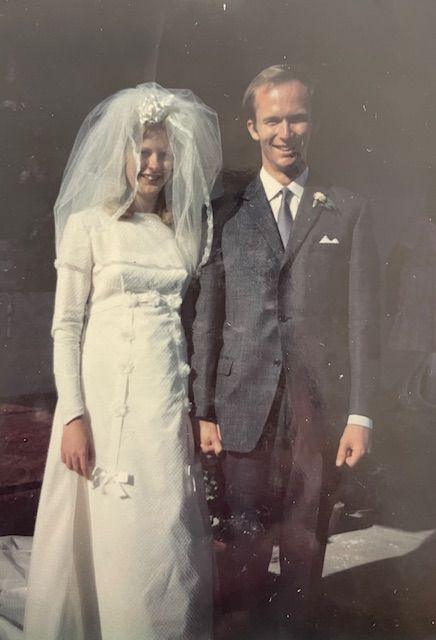 Barbie and Peter Reynolds holding hands and smiling for a portrait picture on their wedding day. Barbie is on the left wearing a wedding dress and veil. Peter is stood next to her in a grey suit