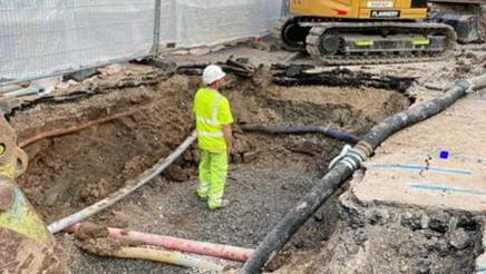 Worker standing in deep pit with soil dug out and pipes exposed. A digger is visible in the background.