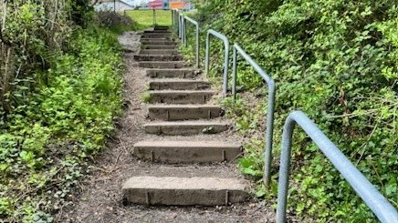 An outdoor footpath made up of a series of concrete steps running up a small incline towards a semi-urban area at the top. On both sides of the steps, there are metal handrails painted grey, running parallel to the path. Green vegetation lines both sides of the path.