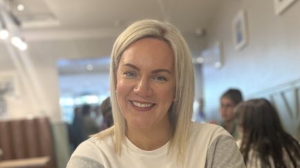 Sarah Harrigan in a restaurant with people in the background. She has blonde hair and is wearing a white and grey sweater. 