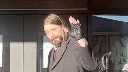 Benjamin Clarke is waving as he walks out of the court building. He is wearing black fingerless gloves and a long, grey coat. He has long brown hair and a beard.