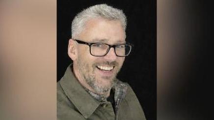 Craig Hurcombe smiles at the camera. He wears a green collared jacket and black rimmed glasses. He has short grey hair and a beard.