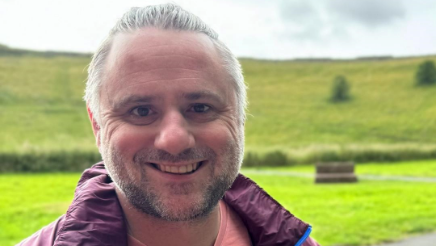 A man wearing a dark red rain coat is standing outdoors with a green field behind him. He has grey hair and a stubbly beard. He is smiling for the photograph.