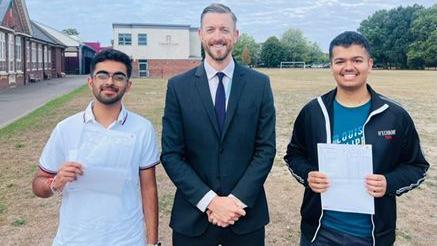 Mr King stands in the middle, with two male students either side of him in school grounds. All three of them are smiling at the camera. The two students are holding their results.