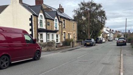 The main street in Ffynnongroyw with houses on either side of the road and cars parked along it.