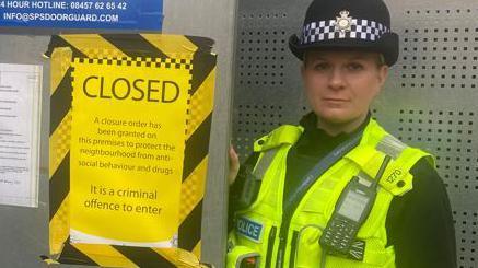 A policewoman wearing a black hat with checked band, black top and yellow vest stands next to a yellow sign saying "CLOSED"